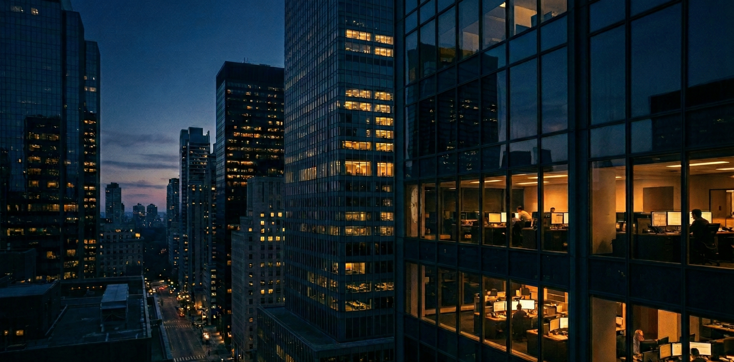 A high-angle photograph looking down an urban canyon of skyscrapers at night. The view is dominated by multi-story, glass-facade office buildings on both the left and right, their grids of windows extending into the distance. Inside several floors of the building on the right, office workers can be seen working late at their desks, with warm indoor lights illuminating detailed scenes of monitors, keyboards, and people at their stations. The buildings on the left are mostly dark, with occasional illuminated windows. Below, a long, straight city avenue stretches into the distance, with the light trails of cars and streetlights visible. A sliver of the dusk sky with a warm sunset glow is visible between the buildings at the very end of the avenue. The perspective creates a sense of towering scale and nocturnal activity.